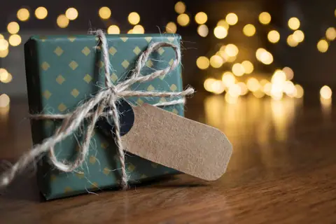 A wrapped gift box with twine beside festive bokeh lights indoors.