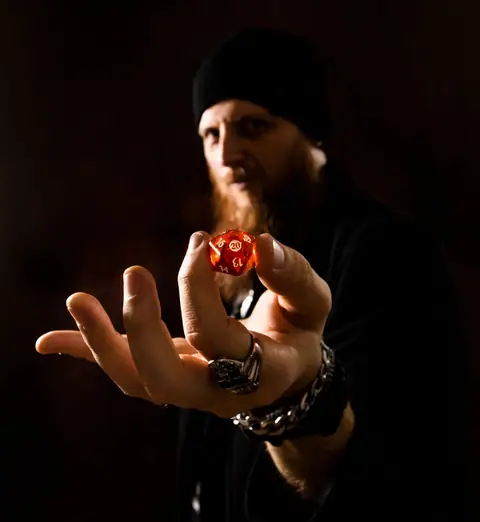 A bearded man holds a red polyhedral dice against a dark background.